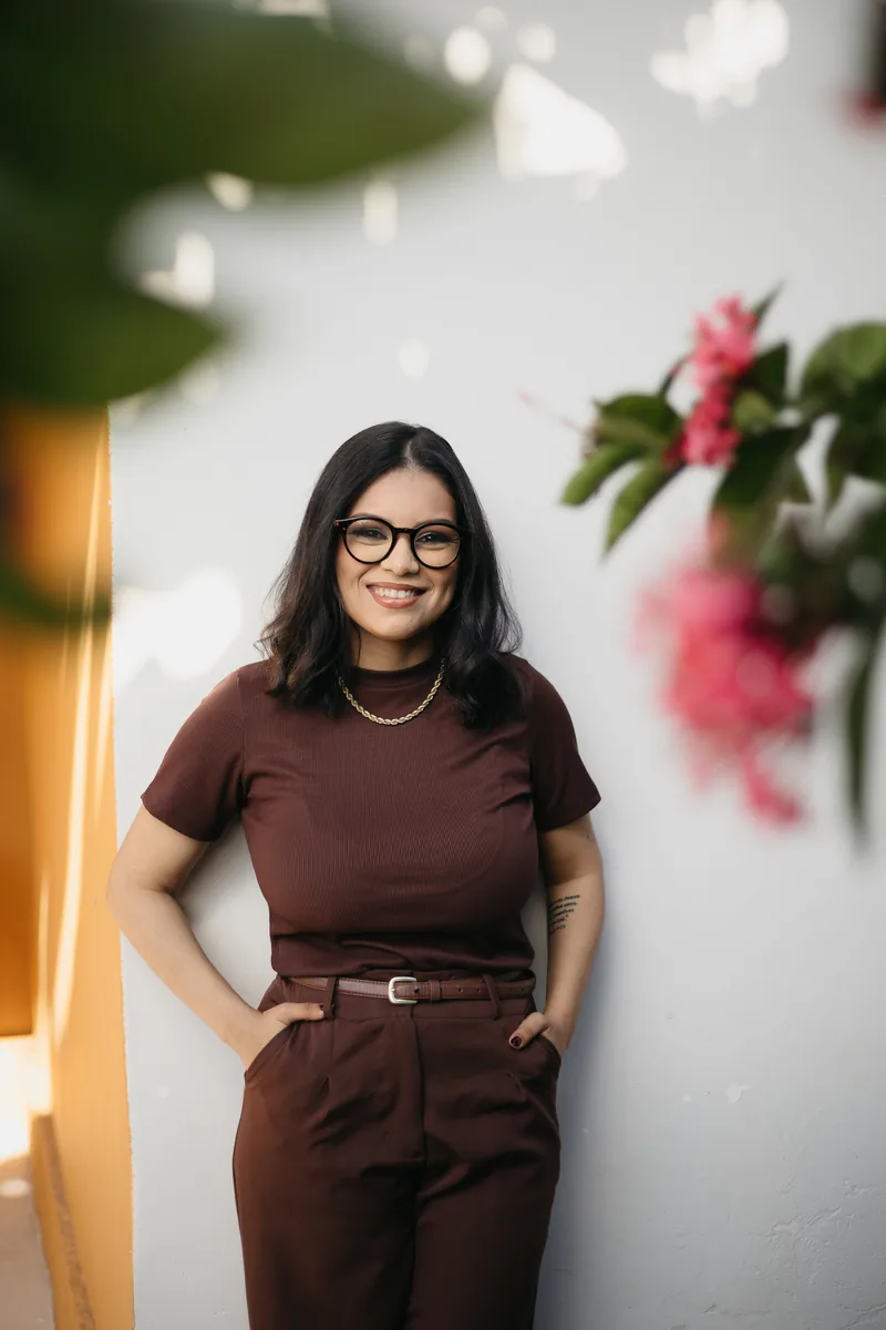 Vitória Gonçalves em pé, sorrindo, diante de uma parede clara com flores